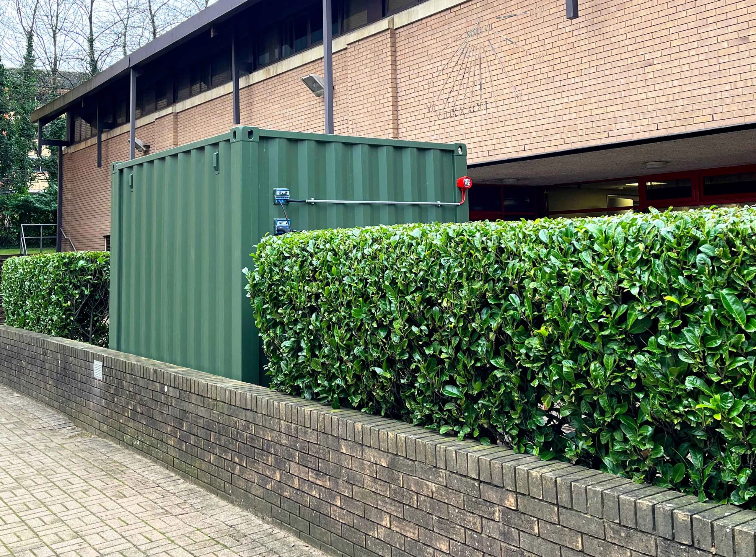 The image shows a green 10-foot metal container located on the Cardiff University campus. It is used for storing and charging batteries. The container is positioned outdoors next to a brick building, surrounded by a neatly trimmed hedge and a low brick wall. The area has a paved walkway in the foreground and a mix of greenery and urban features in the background.