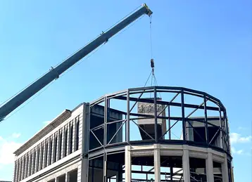 A crane lifts a modified shipping container into place to form the curved front section of the Flatiron Central structure. The container is part of a multi-storey event build and features realistic architectural detailing that mimics stonework. The installation is framed with steel and designed to resemble a traditional building, set against a clear blue sky.