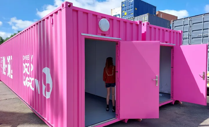 A vivid pink converted shipping container with white branding on the side, featuring two open personnel doors. A person is stepping inside one of the units, revealing a clean, white interior. Stacked shipping containers are visible in the background under a blue sky.
