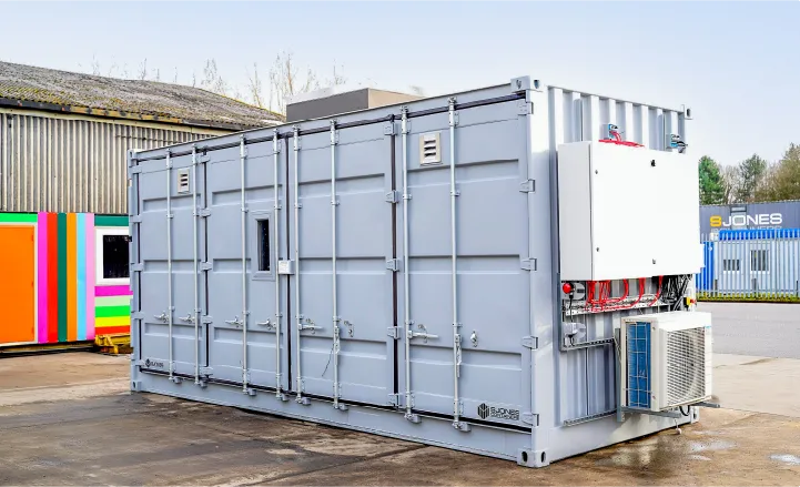 A light grey converted shipping container with external electrical and HVAC components, located outside a warehouse with a brightly coloured unit in the background.