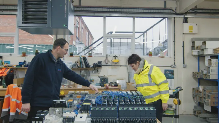 Two people in an industrial workshop examining electrical control panels. One person, wearing a dark jacket and glasses, is pointing at the equipment, while the other, wearing a high-visibility yellow jacket, listens attentively. The workspace contains shelves, tools, and various electrical components, with large windows allowing natural light to enter.