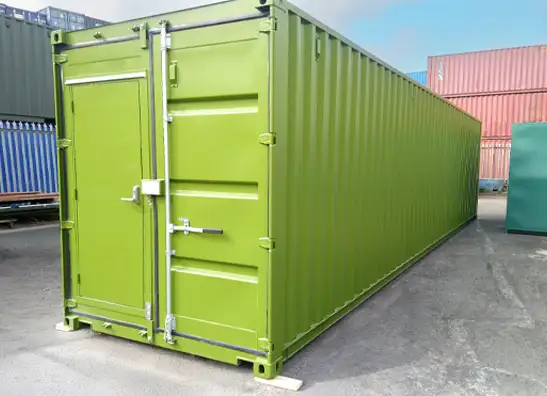 A long green shipping container with a personnel access door integrated into the main double doors, situated on a paved surface with other containers visible in the background.