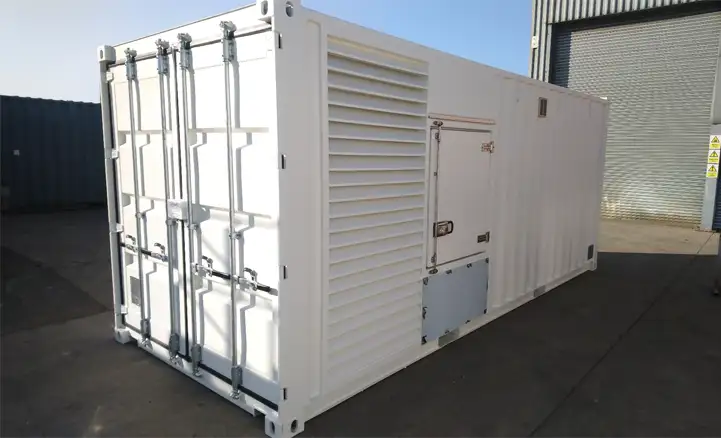 A white shipping container modified to house a generator, featuring large ventilation louvres, access panels, and reinforced doors. It is positioned on a concrete surface outside an industrial building with a roller shutter door. The container is clean and designed for secure, enclosed operation of power equipment.