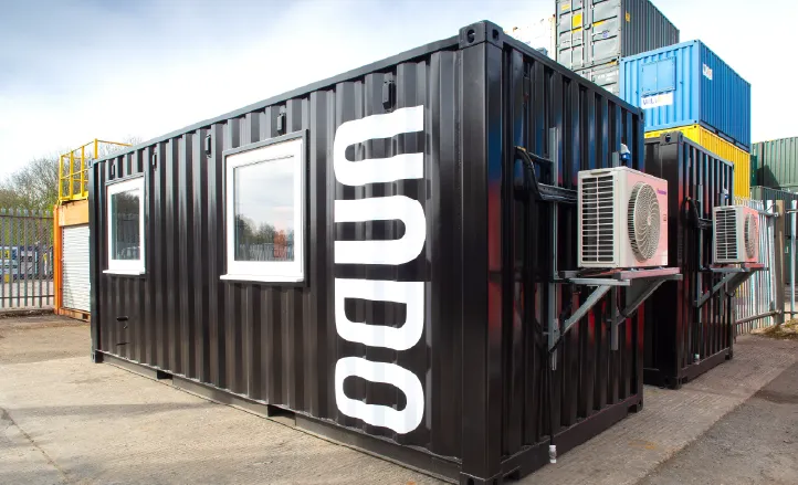 A black converted shipping container featuring large white "UNDO" branding vertically along the side, fitted with windows and external air conditioning units, situated in a yard with other stacked containers.