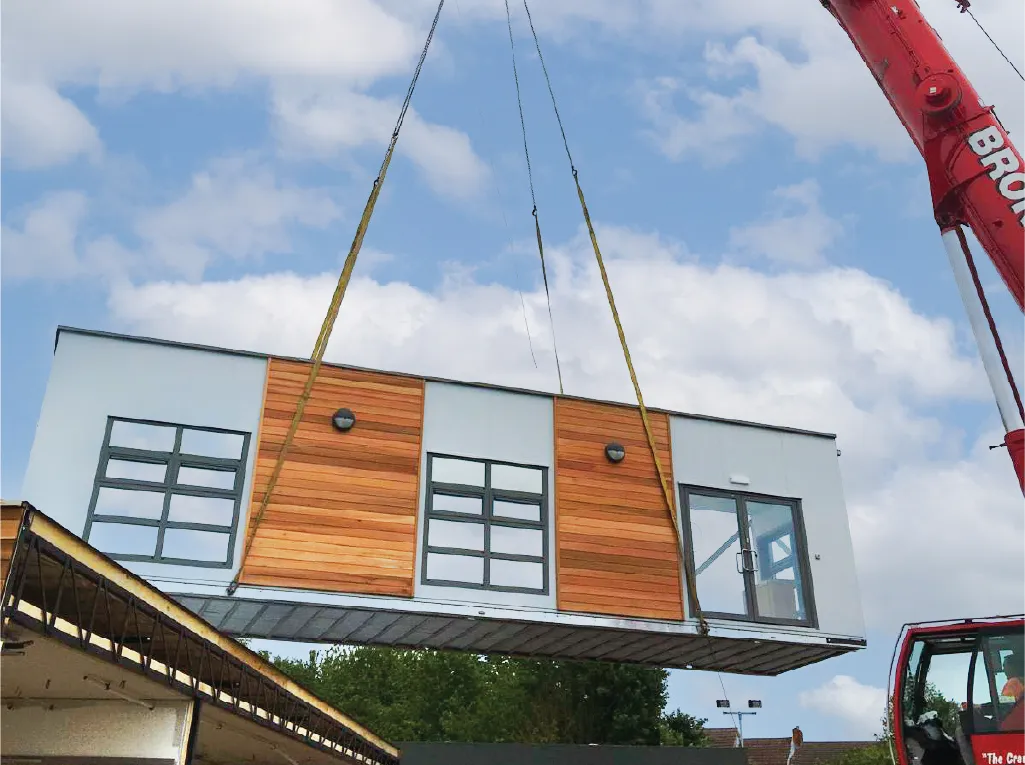 A large crane lifting a modular building section with wooden cladding and large windows into place. The sky is partly cloudy, and the modular unit appears to be part of a larger construction project.