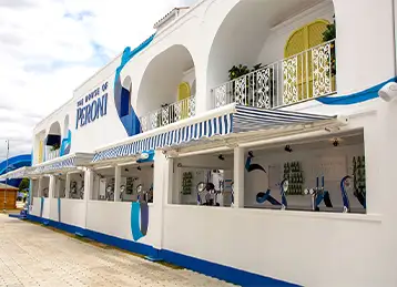 A completed section of the House of Peroni installation, constructed from modified shipping containers. The white façade features arched balconies, decorative railings, yellow doors, and blue artistic accents, creating a Mediterranean-inspired look. An open bar area with striped awnings and Peroni branding is visible, offering service to guests in a stylish, festival-style setting.