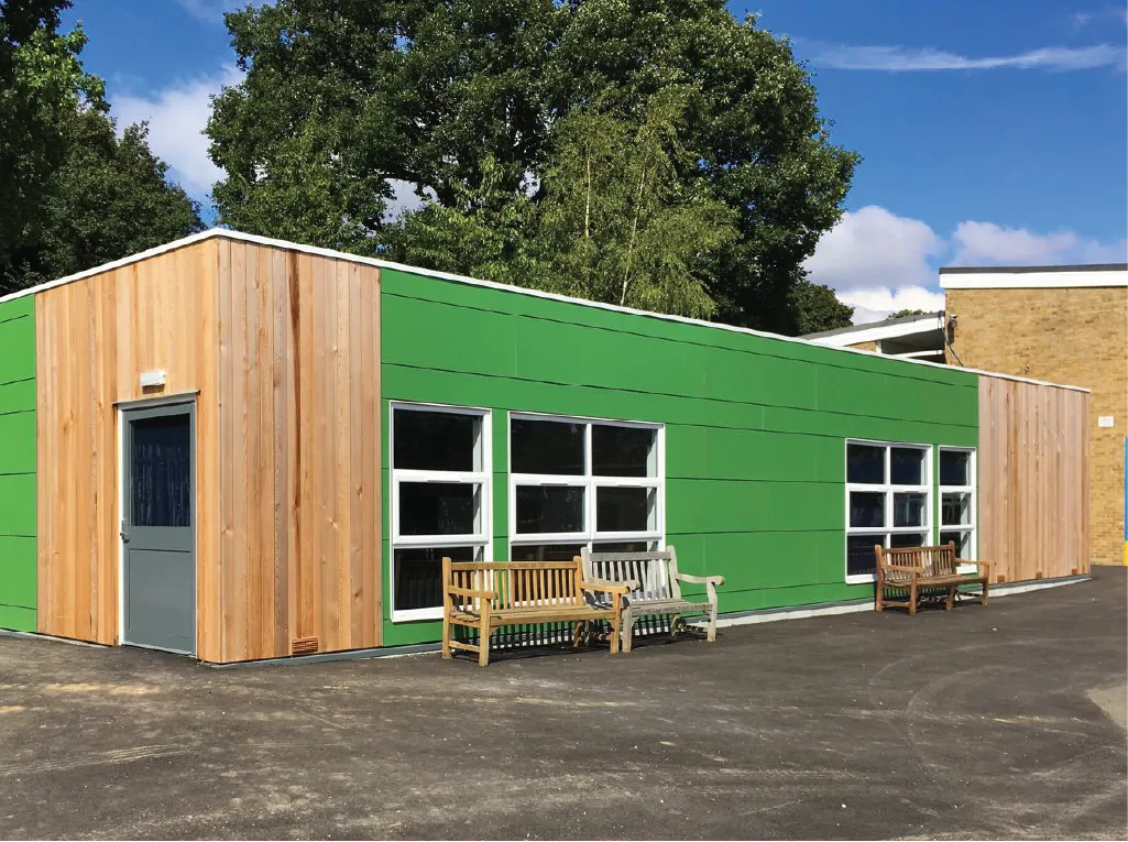 A modern modular building with a green exterior and wooden panel accents. Large windows allow natural light inside, and wooden benches are placed outside on a paved area, surrounded by lush greenery and trees.