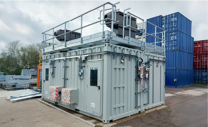A modified shipping container for Battery Testing at Warwick Manufacturing Group painted light grey, equipped with external control panels, ventilation units, and a rooftop platform with metal railings. The container is situated in an industrial yard with stacks of standard blue and red shipping containers in the background.