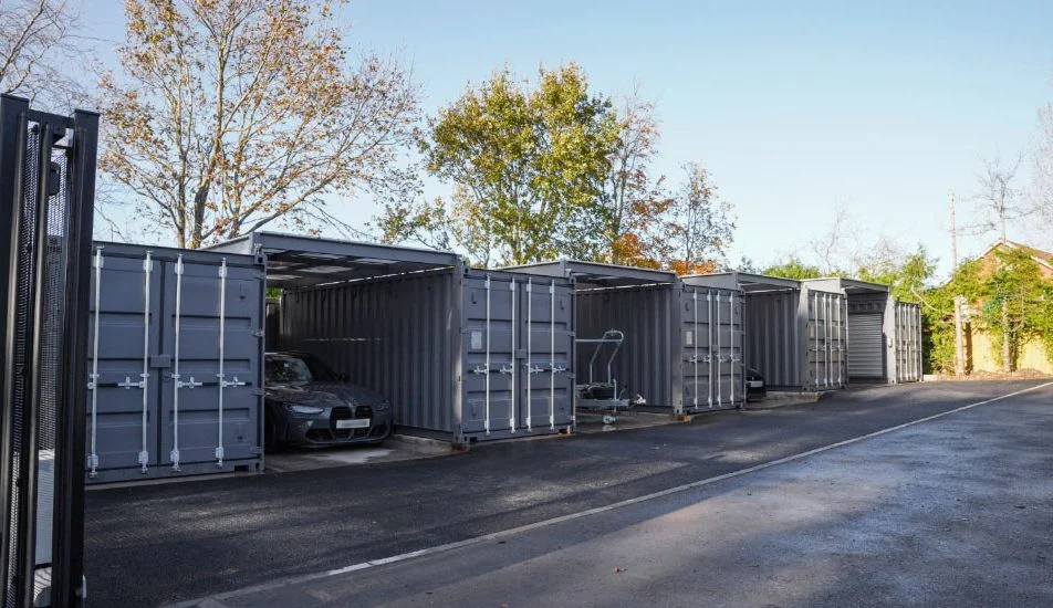A row of modified shipping containers repurposed as storage units is lined up along a paved area, with canopies connecting them to provide additional covered space. The surrounding area features trees with autumn-colored leaves, and a gated entrance is visible in the foreground. The setting is an outdoor facility with a clean and organised layout.