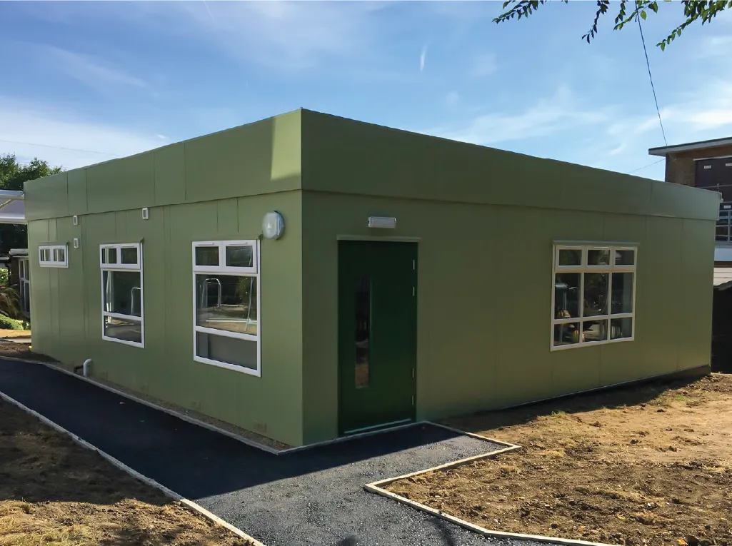 A compact, green modular building with a flat roof, white-framed windows, and a green entrance door. A paved walkway leads up to the entrance, with a dirt-covered landscape surrounding the structure.