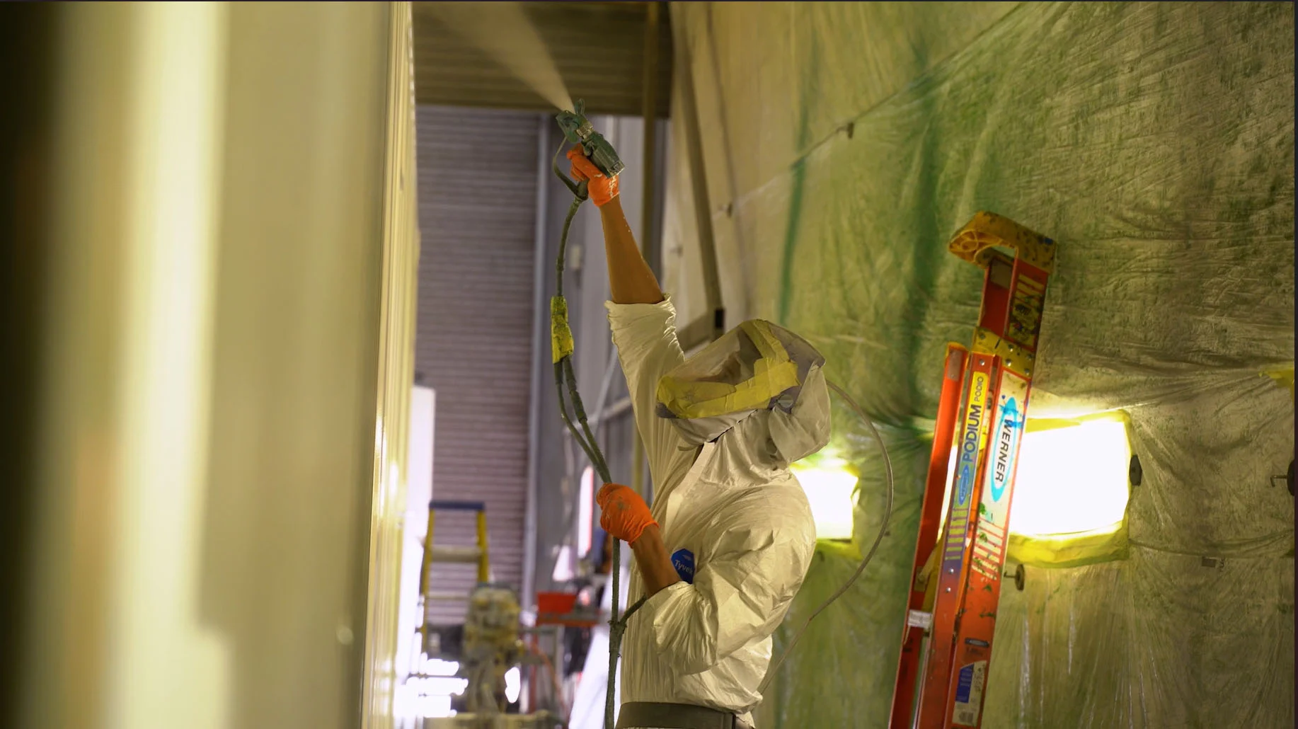 A worker in protective gear, including a full-body suit, gloves, and a hood with a face covering, is spray-painting a large shipping container inside a paint booth. The worker is holding a spray gun high above their head, applying an even coat of paint. The surrounding area is covered in protective plastic sheeting, with bright lights illuminating the workspace. A red ladder and various equipment are visible in the background.