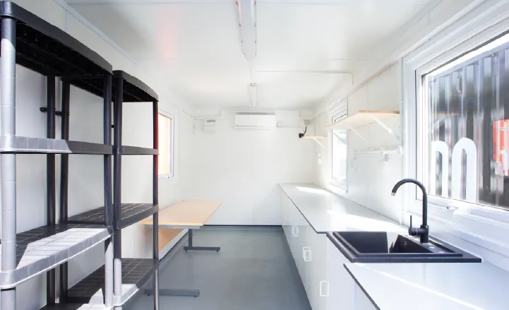 Interior of a converted shipping container with a clean, white fit-out, featuring worktops, a black sink, shelving units, and wall-mounted air conditioning, with natural light streaming through large windows.