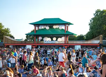 A large crowd gathers in front of the completed Sakura bar installation at an outdoor event. The Japanese-inspired structure features green pagoda-style roofs, red pillars, and hanging white lanterns, built atop a base of modified black shipping containers. The festive scene takes place on a sunny day, with people enjoying the atmosphere and relaxing on the grass.