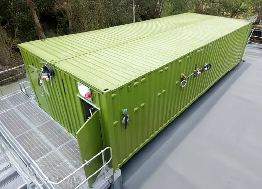 A large green shipping container converted into a switchgear enclosure for CEMA, positioned on a raised platform with surrounding railings. Electrical cables and fittings are visible on the exterior, and the unit is situated at an industrial site with construction machinery in the background.