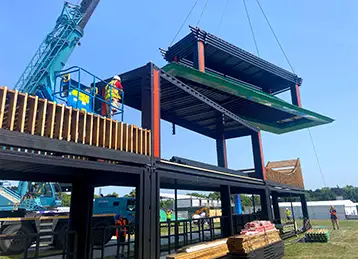 Construction of the Sakura event structure in progress, using modified black shipping containers as the base. A crane lifts a green roof section into place, supported by red vertical beams. Workers in high-visibility clothing stand on a platform to guide the installation. The scene takes place on a grassy field under a clear blue sky.