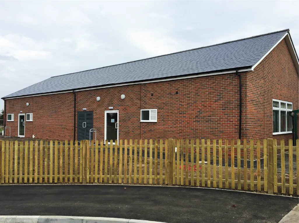 A large, single-story brick modular building with a pitched roof, multiple doors, and windows. A wooden picket fence encloses the area, with a paved driveway in the foreground under an overcast sky.