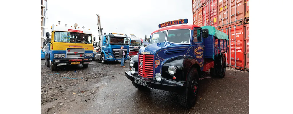 A collection of vintage and classic trucks is parked in an industrial yard with shipping containers in the background. A restored blue and black truck with "Bartlett’s" branding is prominently featured in the foreground, while other colorful classic trucks, including a yellow and blue one, are visible behind it. A man is seen walking among the trucks.