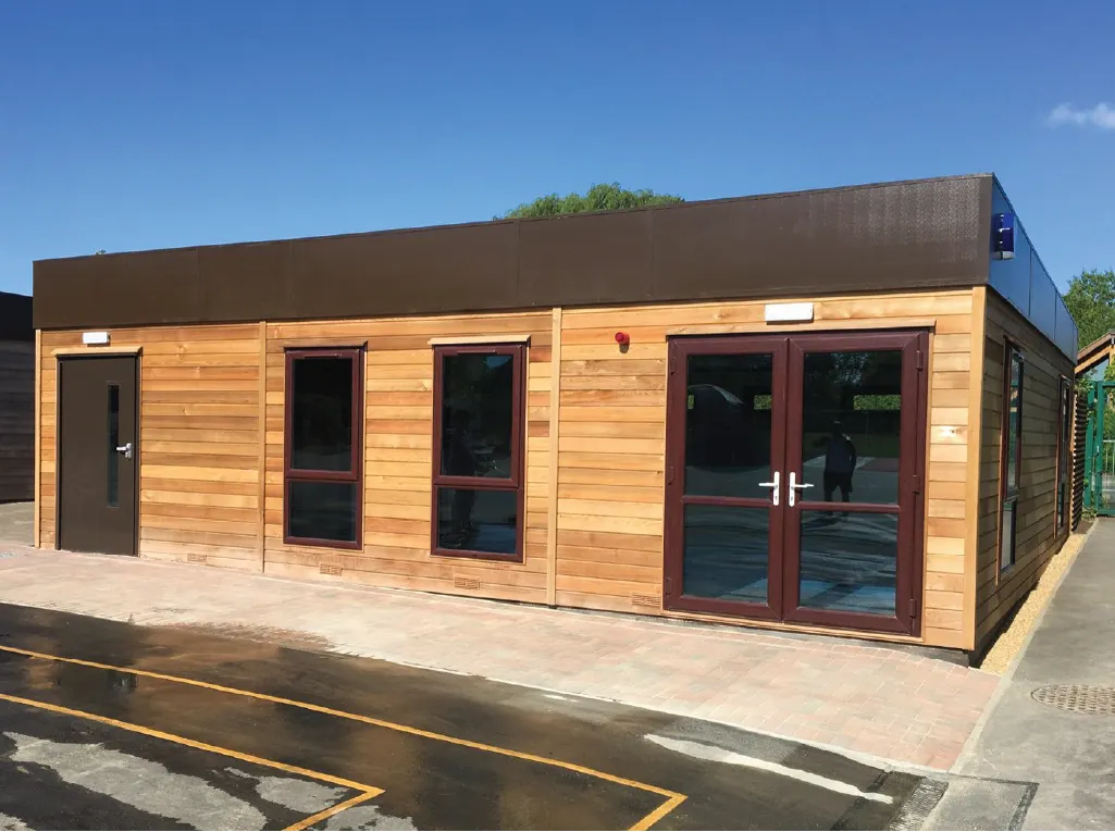 A contemporary modular building with a wooden-clad exterior, brown trim, and large glass doors. The structure is set on a paved area with parking markings, reflecting a bright blue sky.