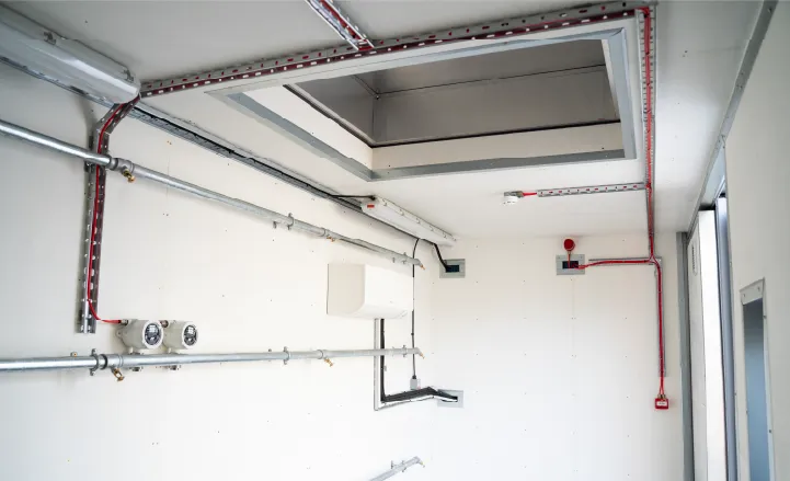 Close-up of the interior ceiling of a converted container, showing ventilation ducting, a roof blast panel, and cable trays arranged neatly along a white wall.
