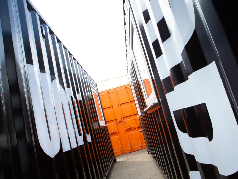 View down the gap between two black shipping containers with large white "UNDO" branding, leading to a striking orange container at the far end.