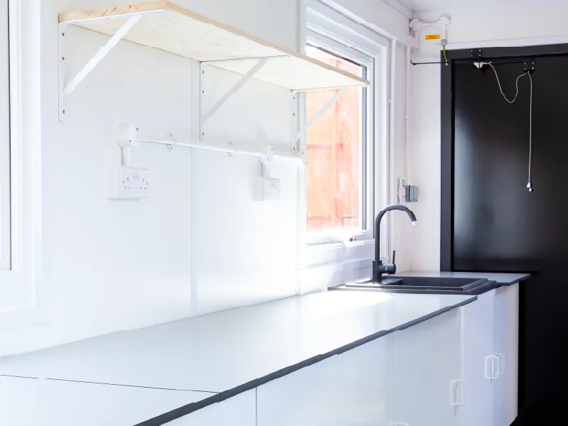 Bright, clean interior of a container conversion showing a black sink and tap, white worktops, under-counter storage, wall-mounted sockets, and open wooden shelving.