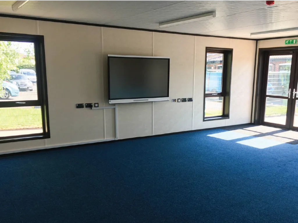 A modern modular classroom interior featuring a mounted interactive screen on the wall, blue carpeted flooring, and large windows allowing natural light to enter. The room is empty, with glass doors leading outside.
