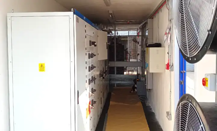 Switchgear equipment installed inside a converted shipping container by S Jones Containers, featuring electrical control panels on the left and cooling fans on the right, with cable management and safety features visible throughout.