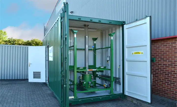 A green shipping container with both doors open reveals industrial water treatment equipment inside, including a vertical assembly of green metal frames and pipework. The container is situated on a paved surface next to a warehouse building with metal siding and a red brick wall. A caution sign is visible on the inside of the container door.