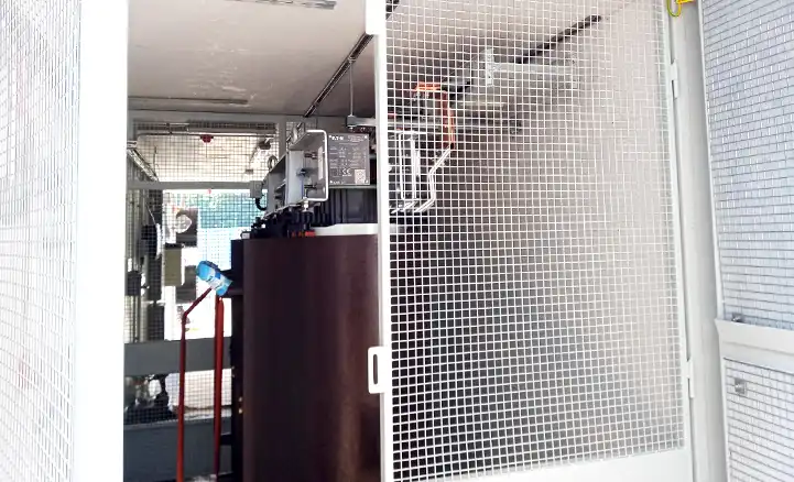 Interior view of a converted shipping container by S Jones Containers, showing electrical equipment securely housed behind a white metal mesh safety cage. Visible components include a large cylindrical unit, likely a transformer, with connected wiring and control devices mounted above.