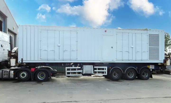Large white containerised generator enclosure mounted on a flatbed trailer, featuring access doors and ventilation panels, ready for transport and deployment to industrial or remote sites.