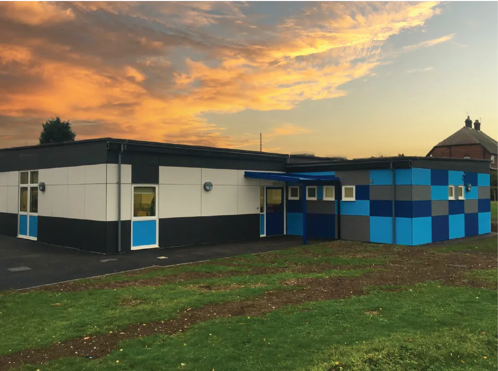 A modular building with a bold exterior design featuring white, black, and blue panels. The setting sun casts a warm glow on the sky, highlighting the unique geometric design of the structure.