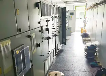 Interior view of a containerised switchgear enclosure featuring rows of electrical control panels and switchgear cabinets along one wall. The space is well-lit with a rubberised anti-slip floor and various tools and equipment temporarily stored along the opposite side.
