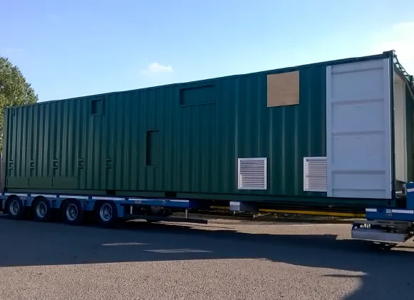 A wide view of a custom green container conversion on a trailer, showing end doors partially open and various ventilation panels and cut-outs on the side.