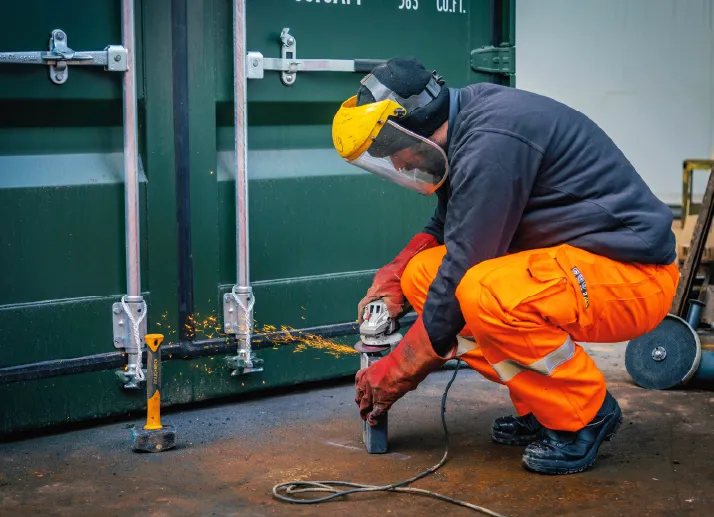 A worker in high-visibility orange trousers and protective gear uses an angle grinder to cut or repair a metal part on a green shipping container, with sparks flying.