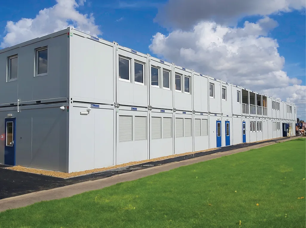 A large, two-story modular building painted white with blue doors and window accents. The structure features multiple windows, some with shutters, and a grassy area in the foreground under a bright blue sky with fluffy clouds.