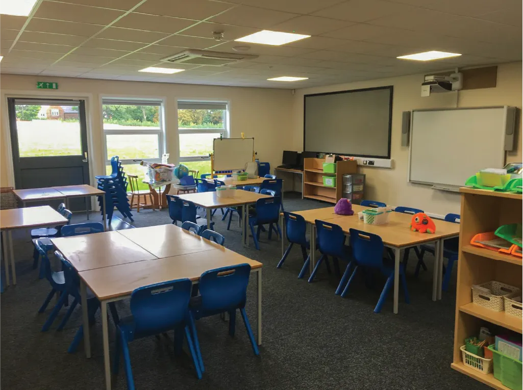 A well-lit modular classroom interior featuring wooden tables, blue chairs, a whiteboard, a projector screen, and educational supplies. Large windows allow natural light to enter, with an exit door leading outside to a grassy area.