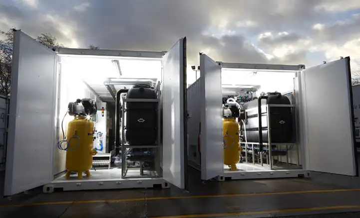 Two shipping containers with their doors open reveal industrial water treatment equipment inside, including large black tanks and bright yellow pressure vessels. The containers are brightly lit from within, contrasting with the dark, cloudy sky in the background. The setup appears to be part of a mobile or modular treatment system.