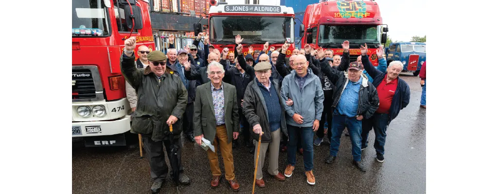 A large group of elderly men, some wearing hats and coats, gather in front of two red ERF trucks with "S. Jones of Aldridge Ltd." branding. Many of them are smiling and raising their hands in celebration. The group appears to be part of a reunion or special event.