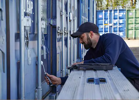 A close-up of a depot worker applying restoration treatment to a shipping container door using a small roller, seated on a platform for easy access.