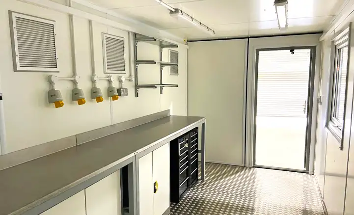 Interior of a converted shipping container workshop with metal workbench, integrated storage drawers, electrical outlets, ventilation panels, and anti-slip flooring, designed for industrial or on-site use.