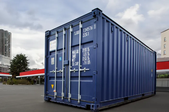 A large blue shipping container with white text displaying weight and capacity information is placed on a paved surface in an urban setting. The container has secure metal locking bars on its doors. In the background, there are tall buildings, trees, and a structure with red and white awnings, possibly a fueling station or parking area. The sky is overcast.