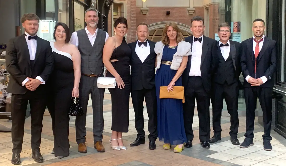 A group of nine people dressed in formal evening wear, posing together and smiling outside the Macdonald Burlington Hotel. The men are mostly wearing tuxedos or suits, and the women are in elegant black and blue dresses with accessories. The setting features a covered walkway with tiled flooring and brick walls in the background, adding a touch of classic architecture to the scene.